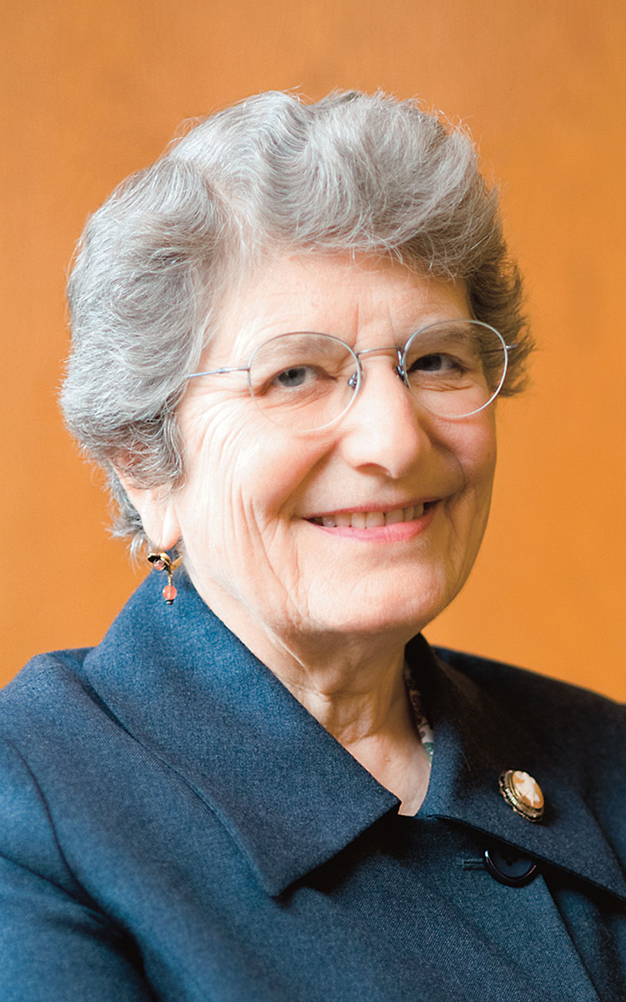 Portrait photo of a smiling Roberta Karmel wearing a navy coat, wireframe glasses, and orange jewelry against an orange background