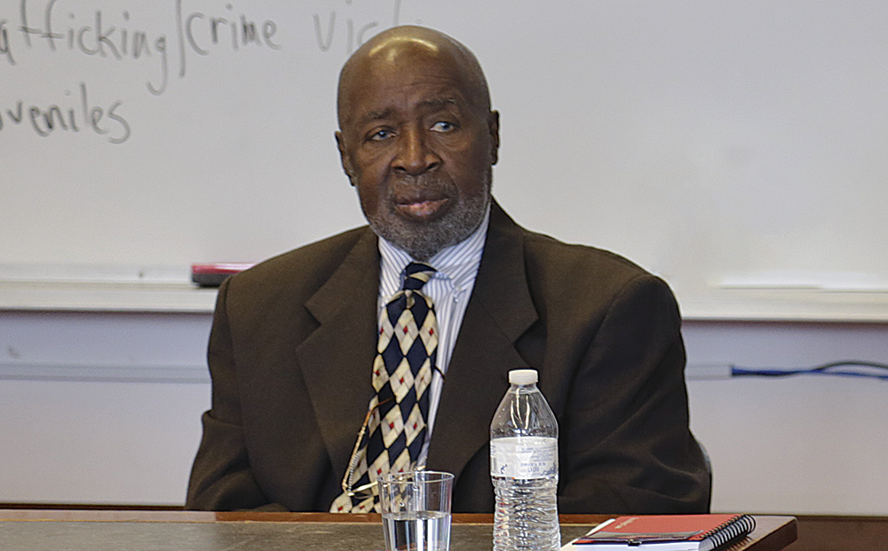 Landscape headshot photograph of Hon. Sterling Johnson Jr. '66 in a dark brown suit, white button-up dress shirt with dark grey lines pattern style, and multi-colored pattern style tie (dark navy blue, tan, red) as he sitting down at a desk with a notebook, glass of water, and plastic water bottle nearby him as Sterling is glancing at something in the foreground